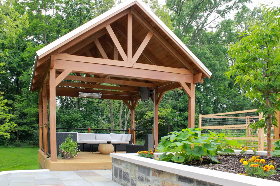 Covered wood pavilion with stone terrace, raised planting bed, flowers, and wooded lawn.